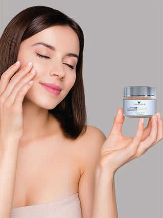 Woman applying cream to her face with a jar of cream in her hand against a gray background