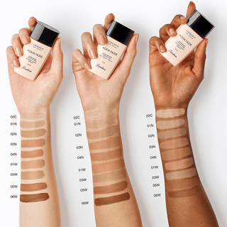 Three hands holding Guerlain foundation bottles with swatches on arms against a white background