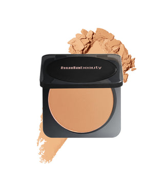 Huda Beauty compact powder with open lid showing product, surrounded by scattered powder on a white background.