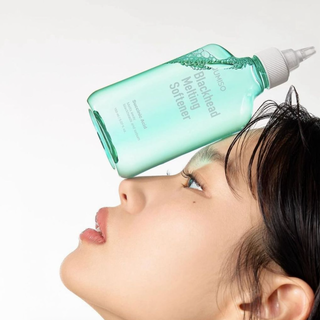 Person applying a green bottle of skincare product to their face against a white background