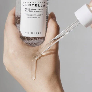 Hand holding a skincare product with a dropper, featuring 'SKIN1004' branding.
